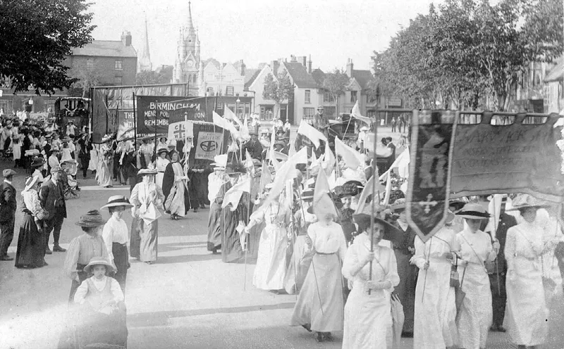Photograph of women in white Edwardian dress marching with banners and flags in a suffragette demonstration, circa 1910